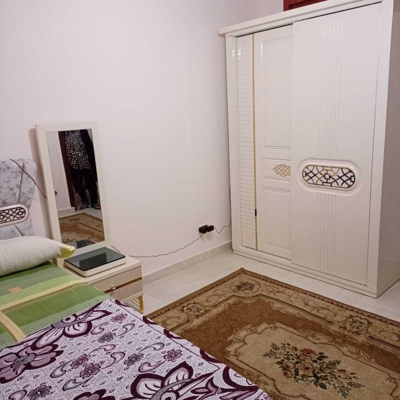 A bedroom with a bed, a bedside table, a mirror, a wardrobe, and a patterned rug.