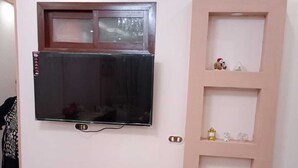 Un televisor montado en la pared con un mueble estante decorativo debajo, que incluye una pequeña figurina y un reloj.