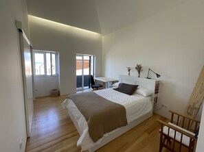 A bedroom with a large bed, a desk, and a chair. There are windows and a view of the outside. - Casa Mar Adentro (Câmara de Lobos)