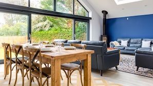 A modern living room with a dining area, featuring a large window, a black sofa, and a wooden dining table. - The Garden House (Wadebridge)