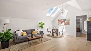 Ein modernes Wohnzimmer mit grauem Sofa, einem Holztisch und einem Essbereich mit rundem Tisch und Stühlen. Es gibt Dachfenster, einen Blumentopf und ein farbenfrohes abstraktes Gemälde an der Wand.