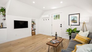 Ein geräumiges Wohnzimmer mit Holzboden, einem Wand-Fernseher, einer Couch mit Kissen und einem Couchtisch mit einer Zeitschrift, sowie einer Tür zu einem anderen Raum.