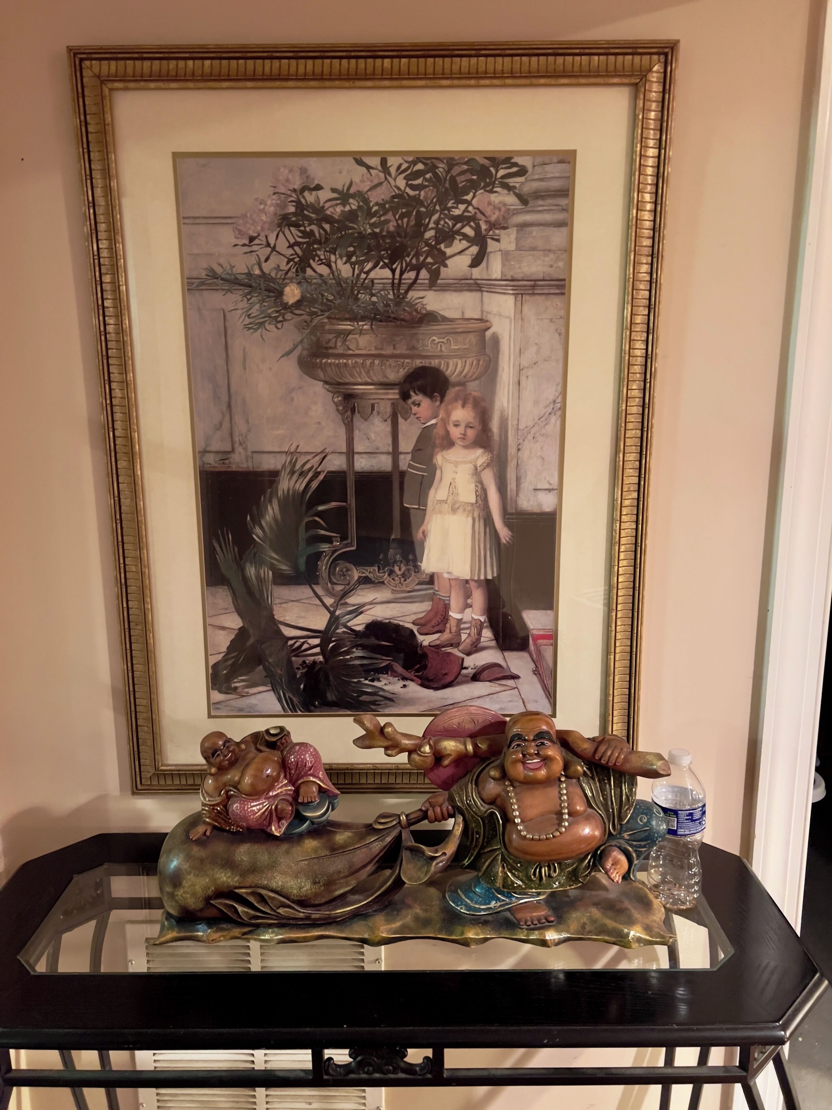A framed black and white photograph of a child and an adult in a room with a marble floor, a large plant, and a decorative table with two figurines.
