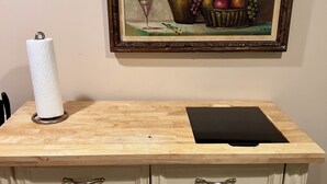 Uma bancada de cozinha com um suporte para toalhas de papel, uma tábua de corte preta e um quadro com uma pintura de natureza morta em cima.