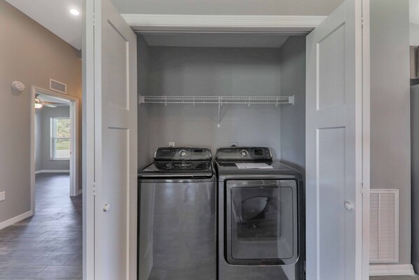 Une buanderie équipée de deux machines à laver et deux sèche-linge superposés, une étagère en métal au-dessus, et un couloir menant à une porte dotée d’une fenêtre.