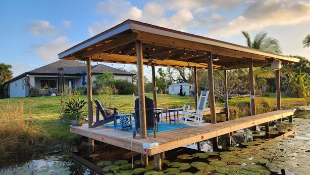 A wooden deck with chairs and a table, surrounded by water and lily pads. -  Lakefront Retreat w/ Dock, Kayaks & Sunset Views – Peaceful Escape! (Lake Placid)