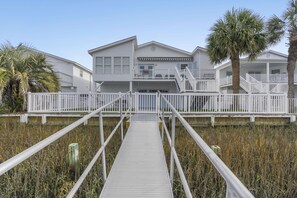 A wooden boardwalk leading to a white building with multiple balconies and a palm tree.