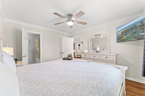 A bedroom with a bed, a ceiling fan, a dresser, a mirror, and a painting of a coastal landscape.