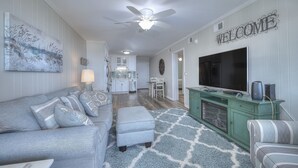 A living room with a grey sofa, ottoman, and striped armchair, a green entertainment unit with a TV, and a 'WELCOME' sign on the wall.