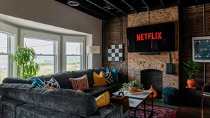 Een moderne woonkamer met een grote grijze bank, een open haard en een televisie bevestigd aan een metselwerkmuur waarop 