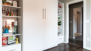 Een kamer met een witte kast, planken met boeken en decoratieve objecten, een houten vloer en een doorgang naar een andere kamer.
