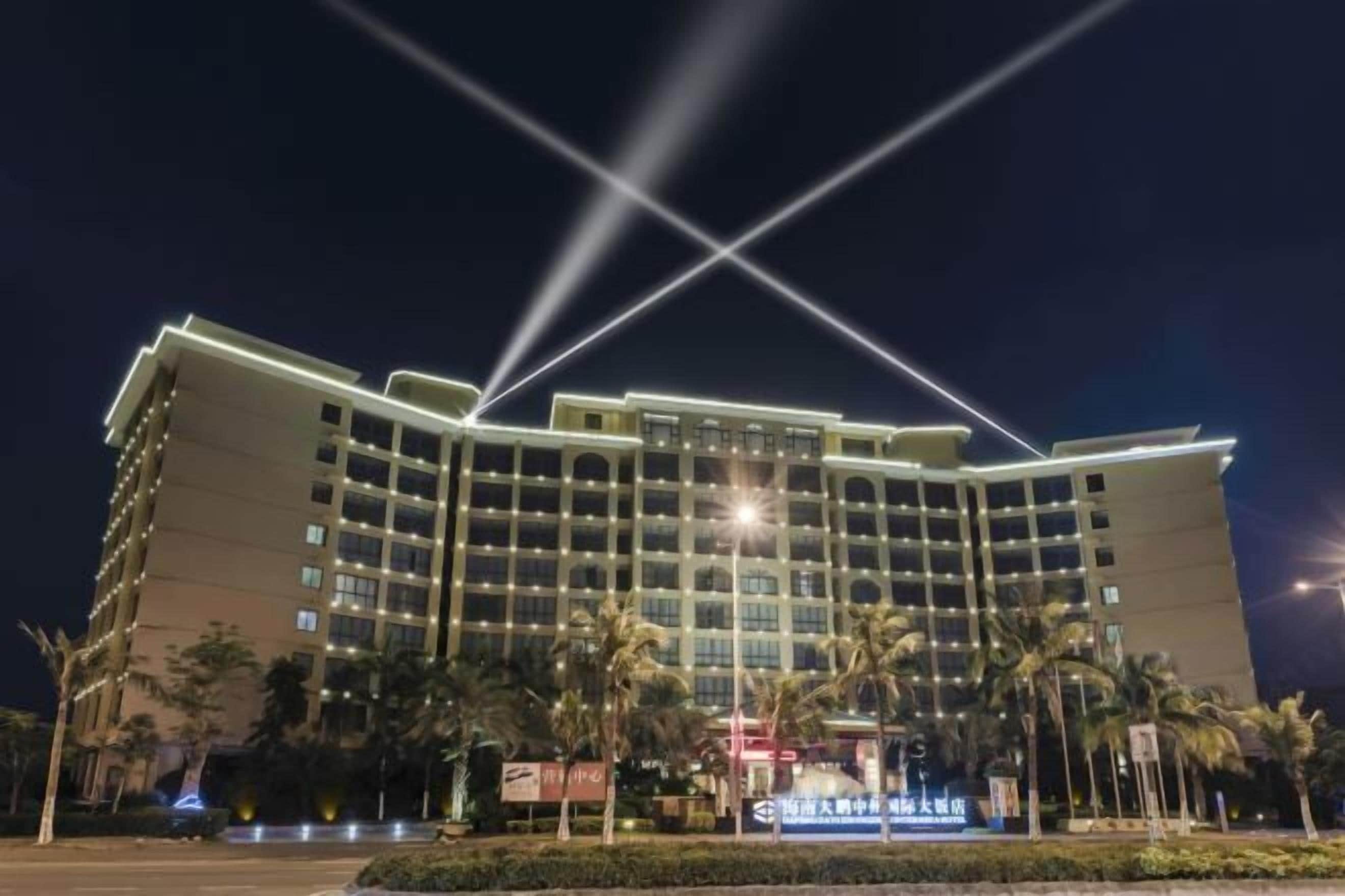 A multi-story hotel with a symmetrical facade, multiple balconies, and a well-lit entrance area with palm trees.
