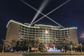 Ein mehrstöckiges Hotel mit einer symmetrischen Fassade, mehreren Balkonen und einem gut beleuchteten Eingangsbereich mit Palmen.
