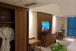 Une chambre d’hôtel avec une armoire en bois, un bureau avec une chaise, une petite table avec une chaise, un téléviseur et une plante en pot.