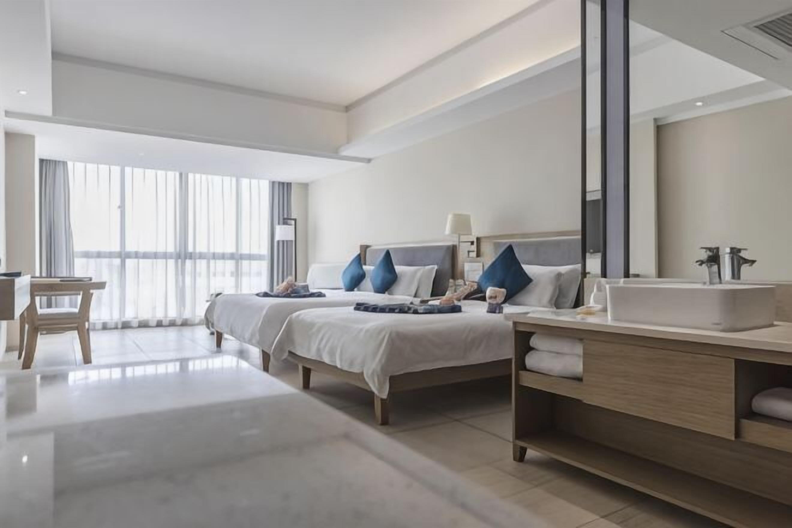 A modern hotel room with two beds, a desk, a vanity with a mirror, and a large window.