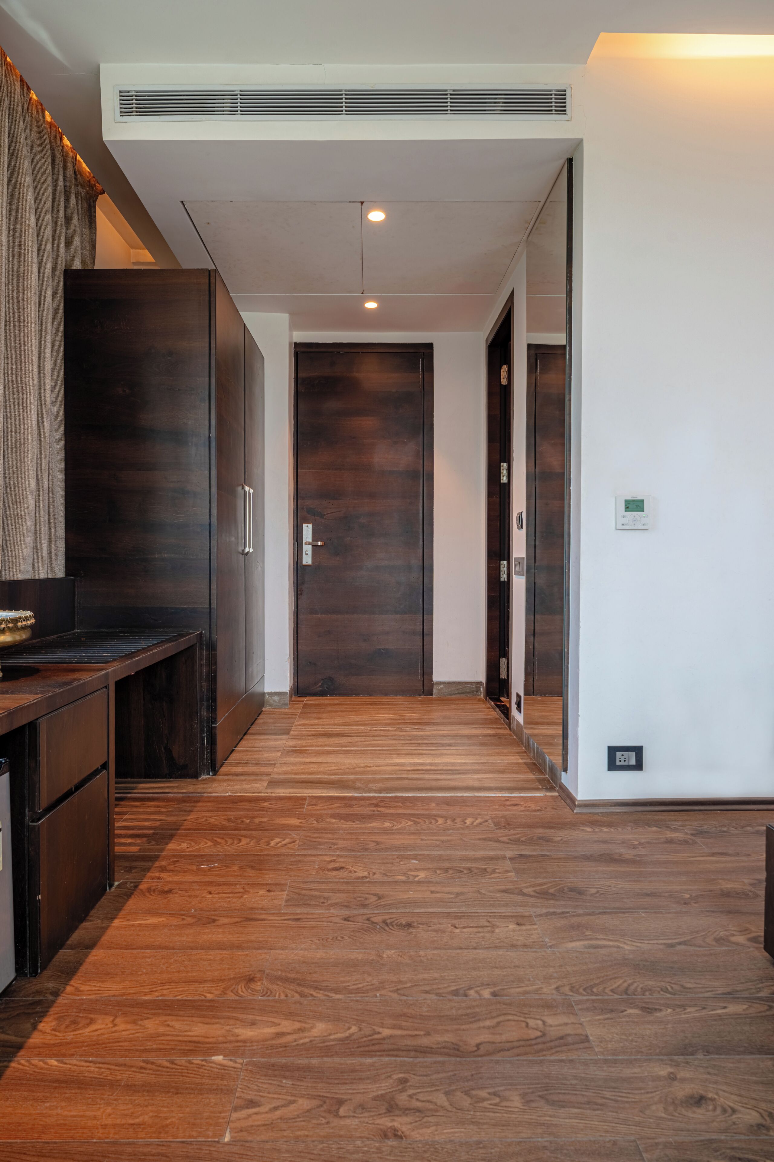 A hallway with a wooden floor, a dark wooden door, and a wall-mounted air vent.