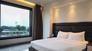 A hotel room with a large window, a bed with white linens, a dark headboard, and a nightstand with a phone. - Econest Hotel (Rampurhat)