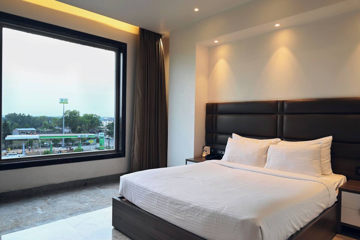 Habitación de hotel con un ventanal grande, una cama con sábanas blancas, cabecera oscura y una mesita de noche con un teléfono.