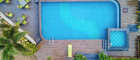 An aerial view of a swimming pool with adjacent lounge area, steps leading to a smaller pool, and surrounding greenery.