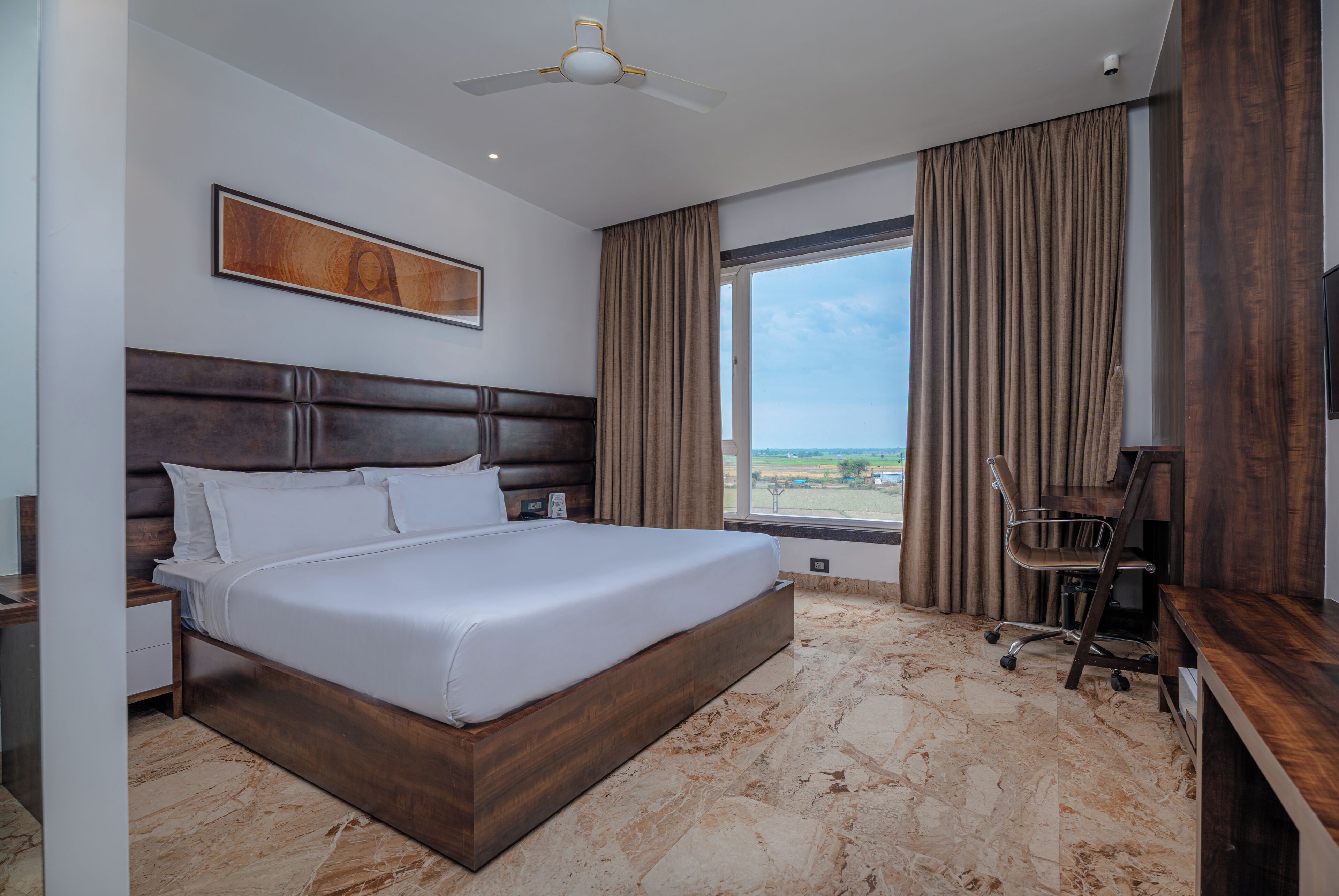 Habitación de hotel con una cama grande, un escritorio, una silla, un ventilador de techo y un ventanal con cortinas.