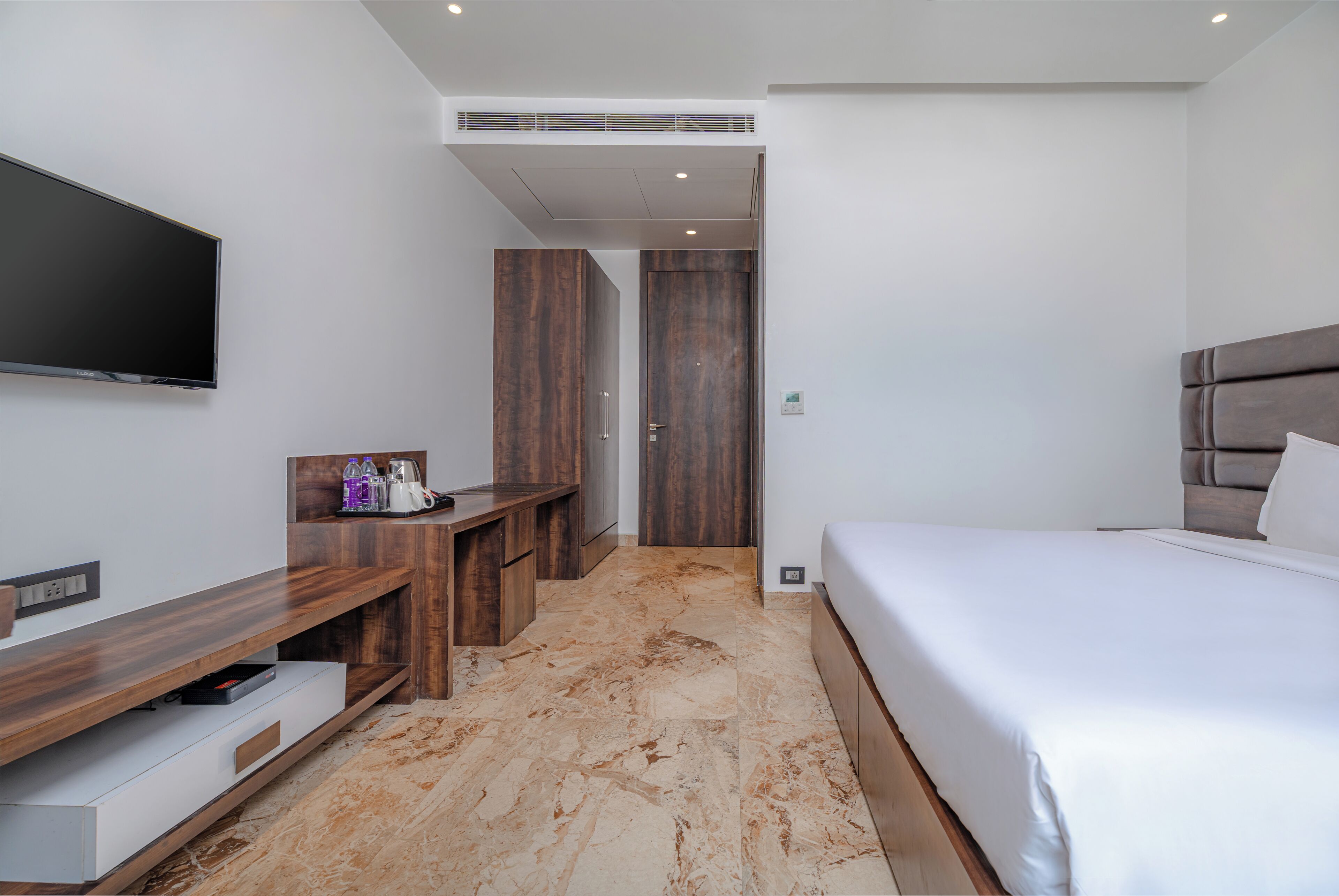A modern hotel room with a flat-screen TV, a wooden desk, a bed, and a bathroom door.