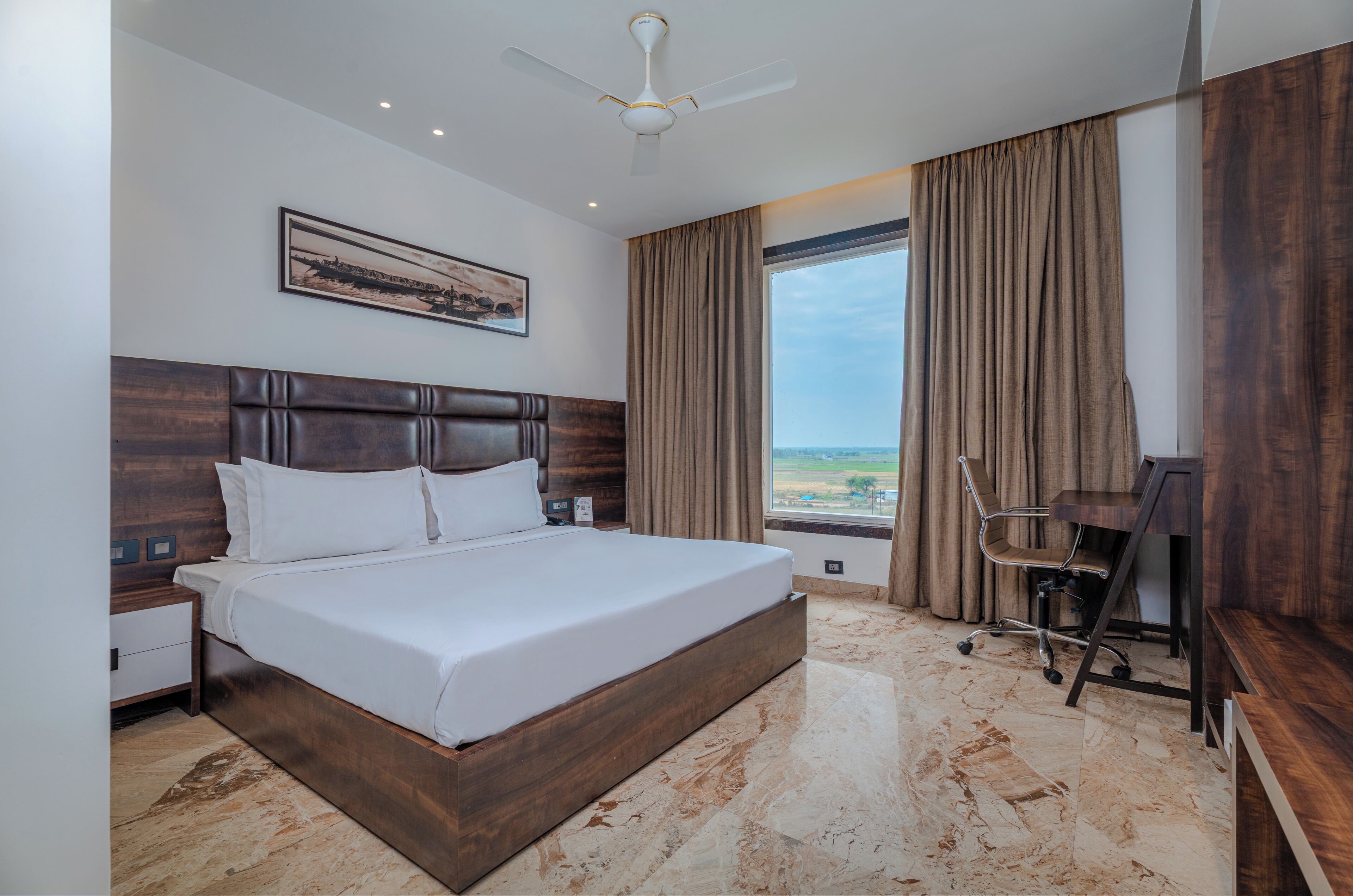 A hotel room with a large bed, a desk, a chair, a ceiling fan, a window with curtains, and a framed picture on the wall.