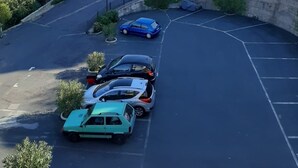 Ein Parkplatz mit Autos, eine gewundene Straße und ein Hügel mit Grünflächen sowie Gebäuden im Hintergrund.