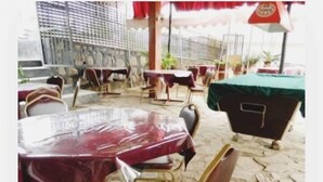Un área de comedor al aire libre con mesas cubiertas de manteles rojos, suelo de piedra y un mantel verde a la derecha.