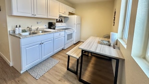 A compact kitchen with white cabinets, a built-in oven, and a coffee maker. There's a small dining table with a woven placemat. - Hot Tub Cozy Cottage in Colorado Springs (Colorado Springs)