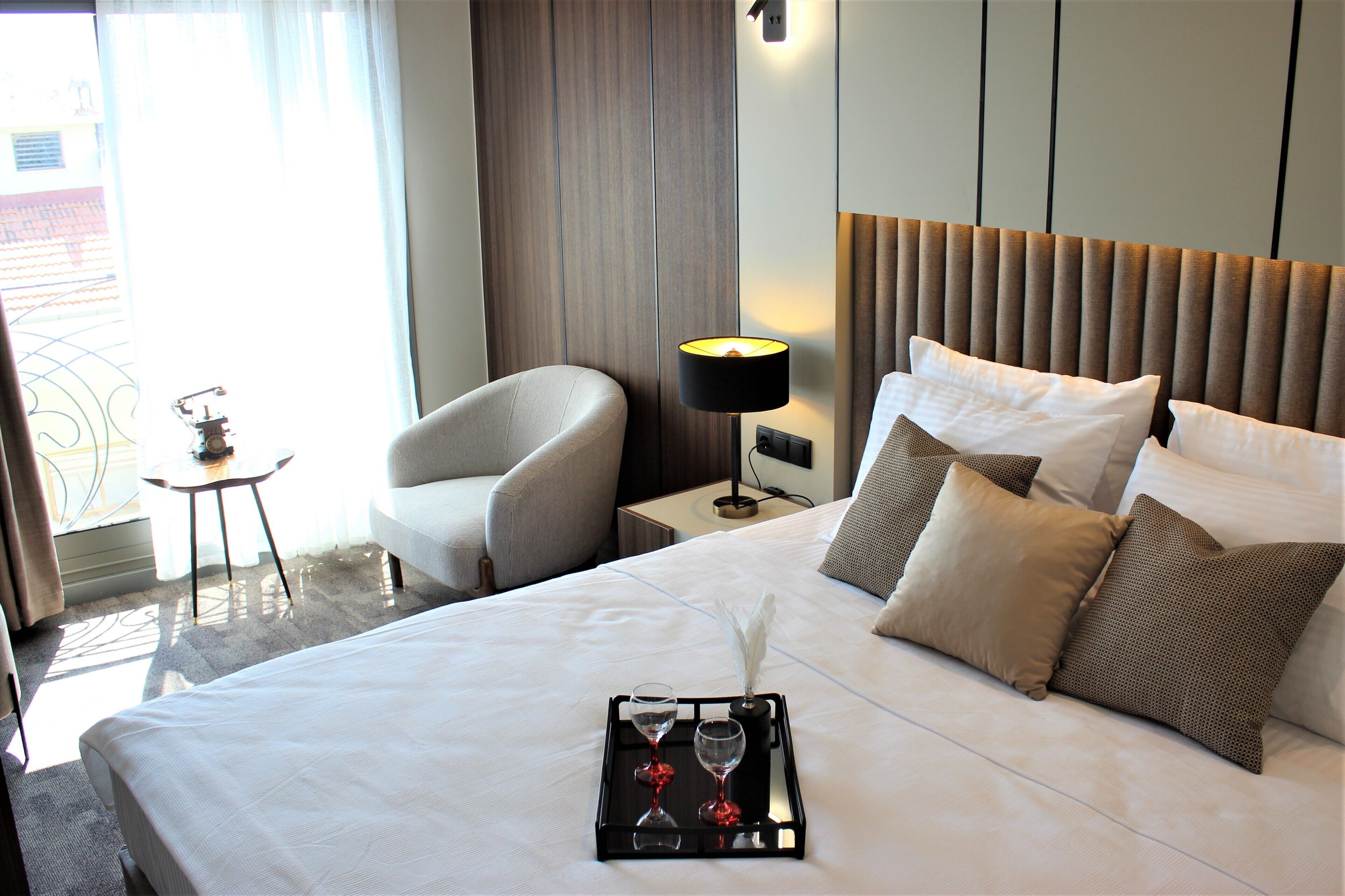 A neatly made bed with a tray of glasses and a feather quill, a bedside lamp, and a chair in a hotel room.