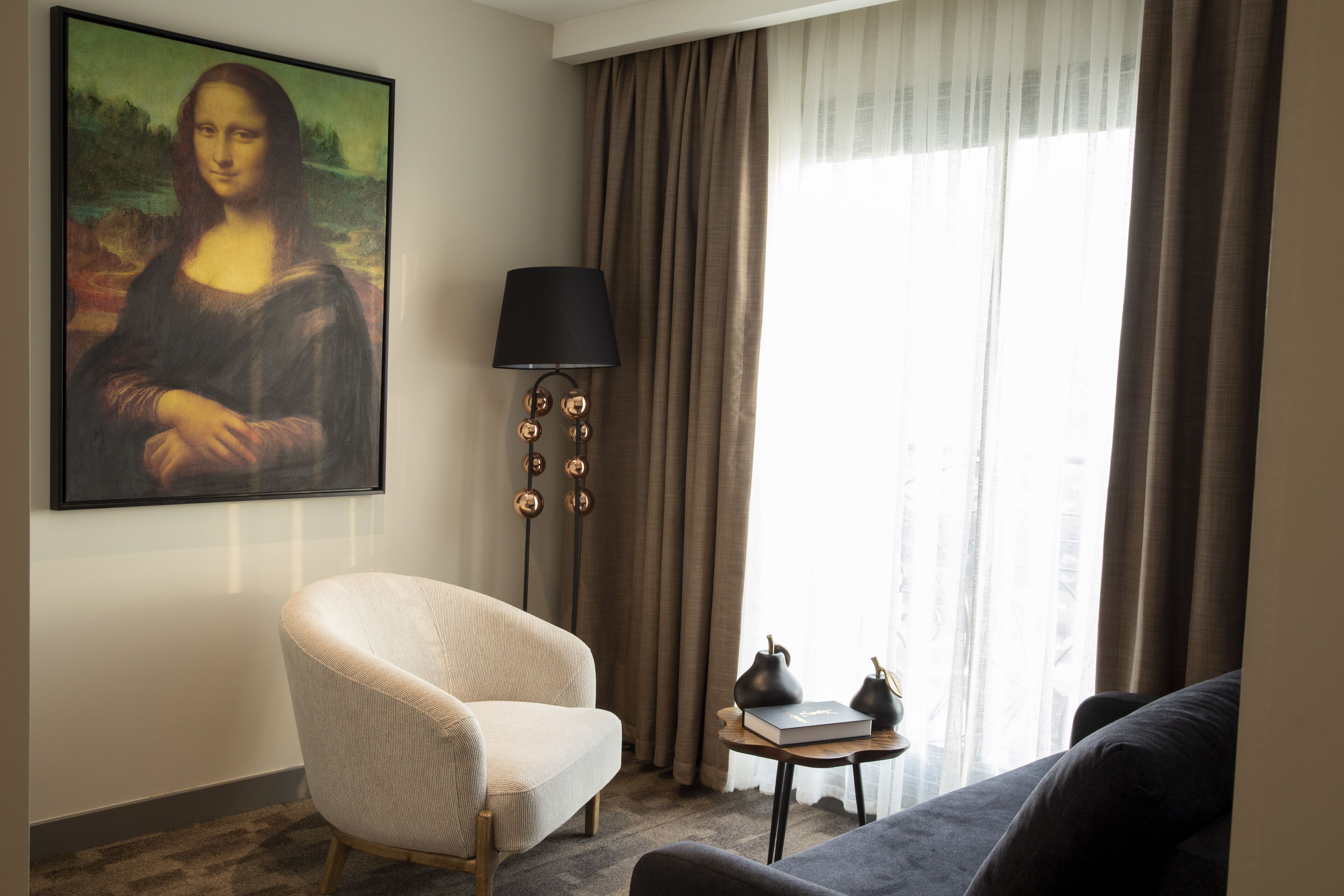 Una habitación con un cuadro grande en la pared, una lámpara, una silla, un sofá y una mesita con libros y una tetera.