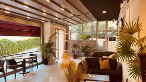 A modern lounge area with a sofa, chairs, and a table. There are potted plants and a view of greenery outside. - Monalisa (Kusadasi)