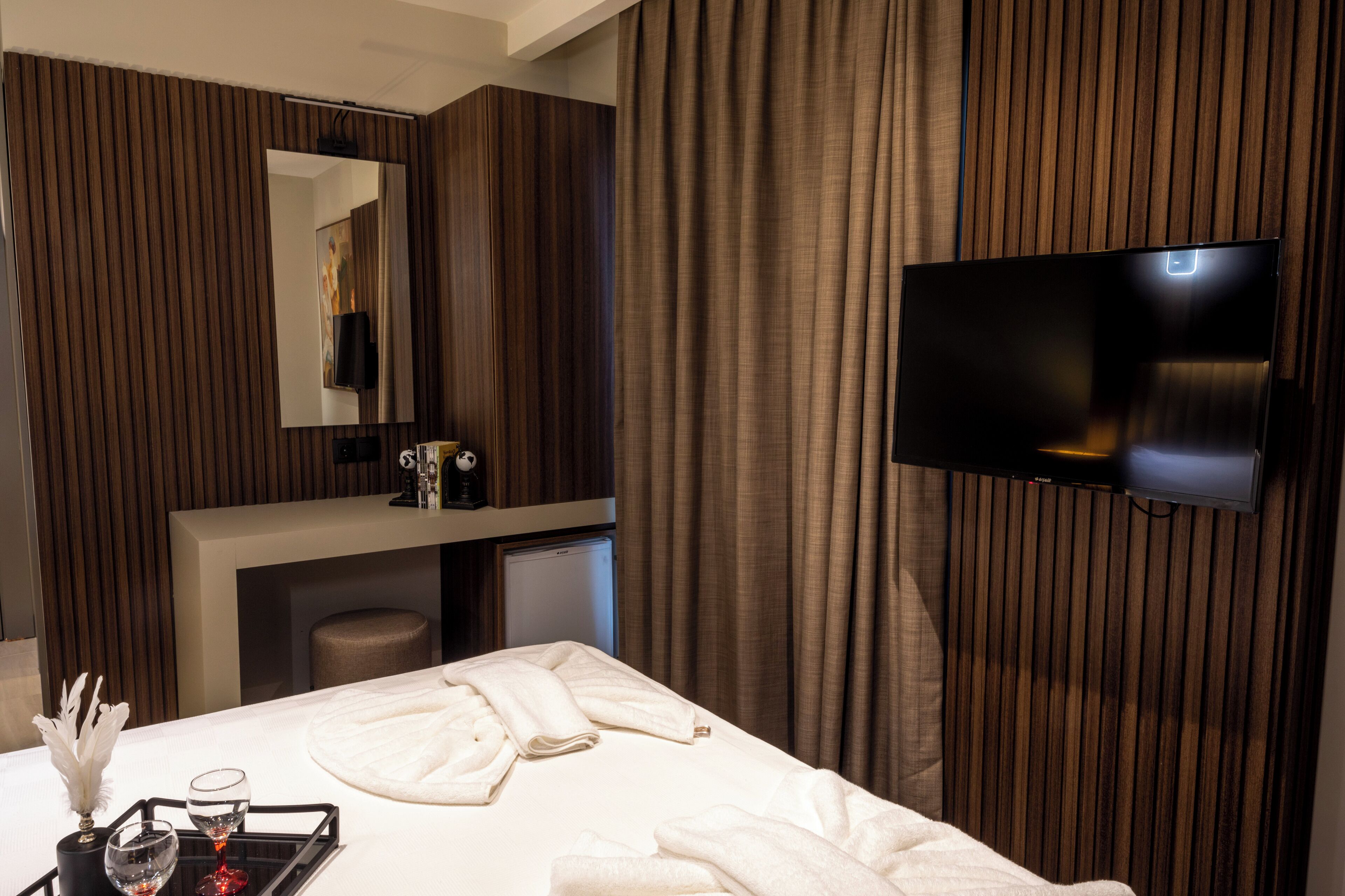 A modern hotel room with a flat-screen TV mounted on the wall, a large bed with white linens, a bedside table with a tray of amenities, and a mirror above the vanity.