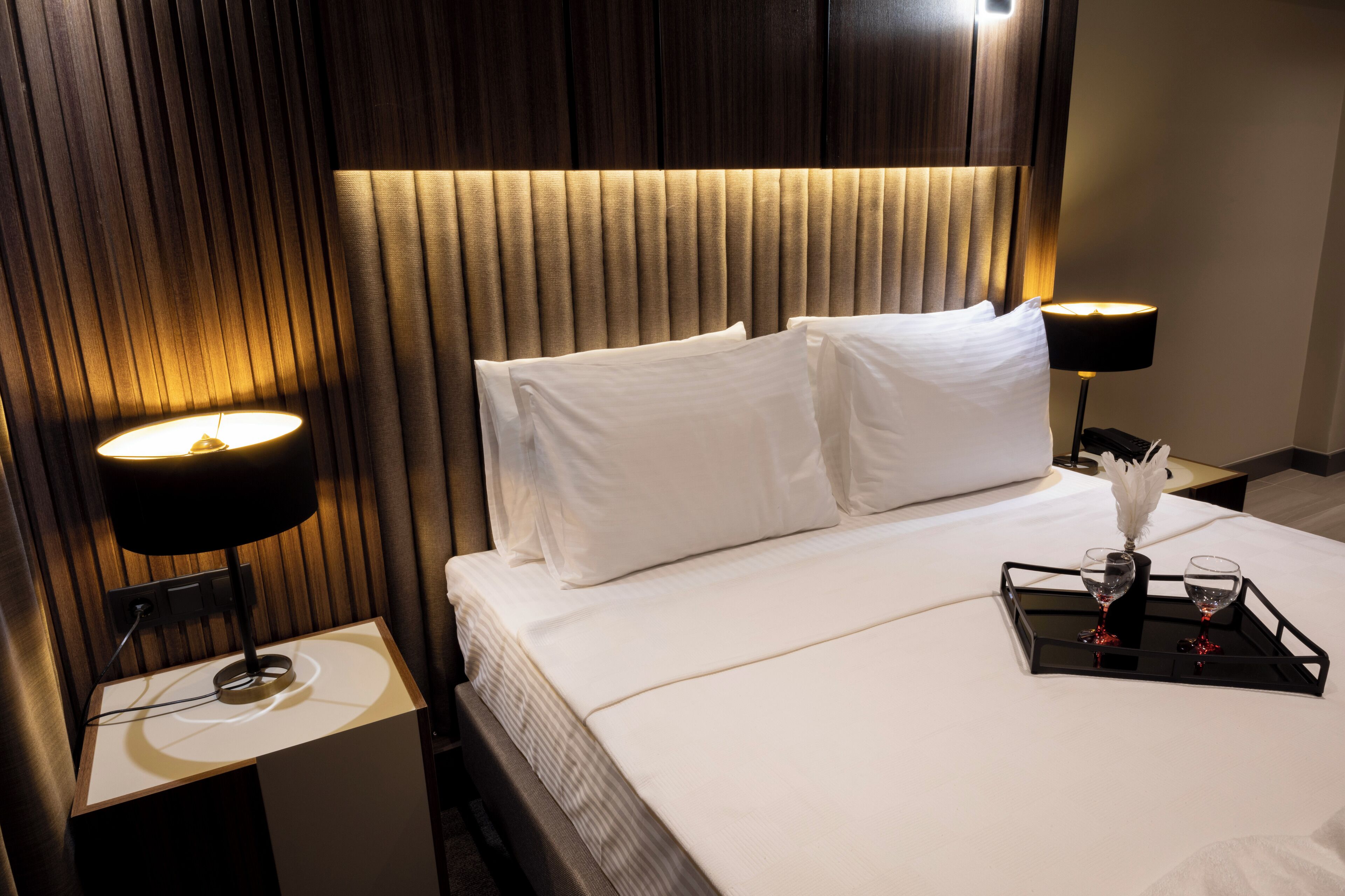 Une chambre d’hôtel avec un lit, deux lampes de chevet, un plateau avec des verres et un vase, et une tête de lit en bois.