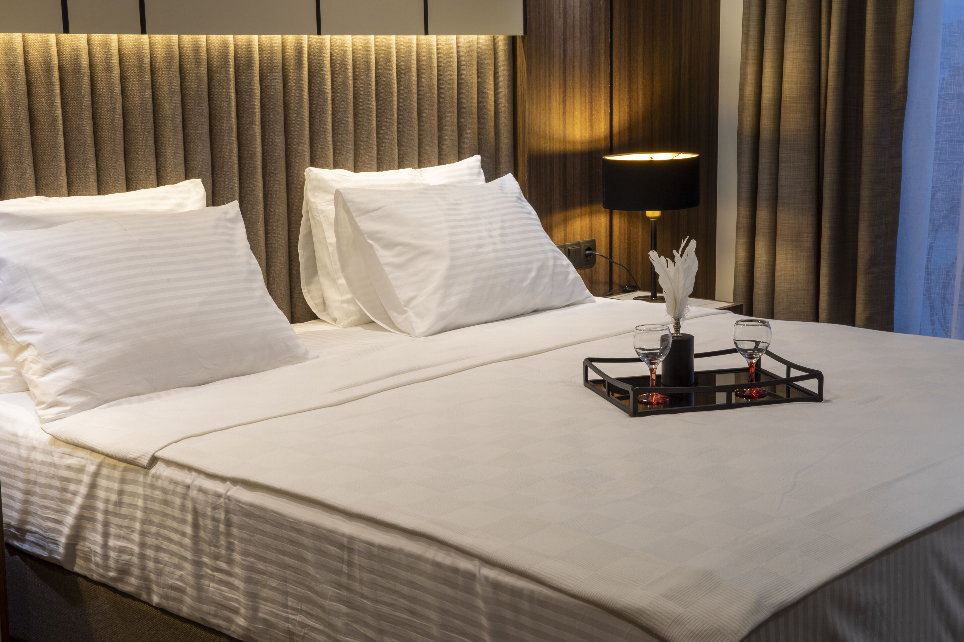 Una cama bien tendida con sábanas y almohadas blancas, una mesita de noche con una lámpara y una bandeja con vasos y un objeto decorativo.