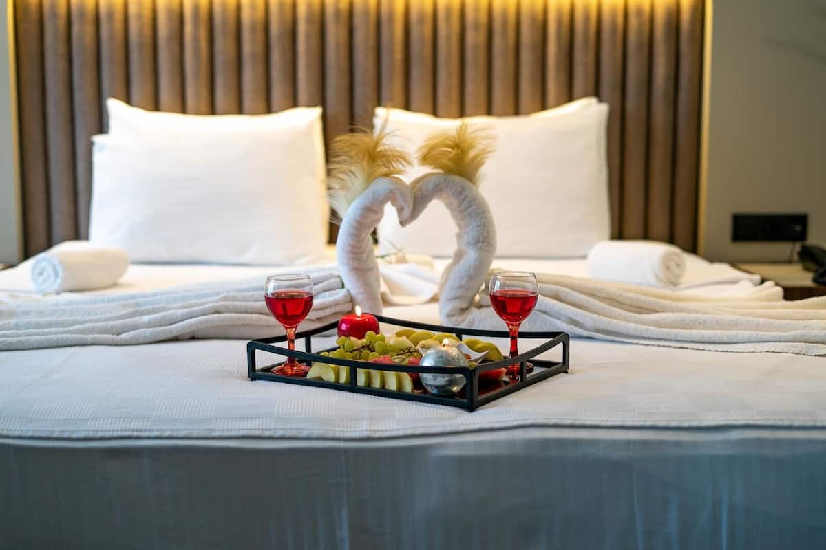 Un lit parfaitement fait, avec du linge de lit blanc, un plateau garni de fruits et deux verres de vin rouge, et un cygne décoratif fait avec des serviettes.