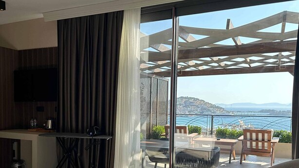 A hotel room with a bed, a desk, a chair, and a balcony with a view of the sea and mountains. - Monalisa (Kusadasi)
