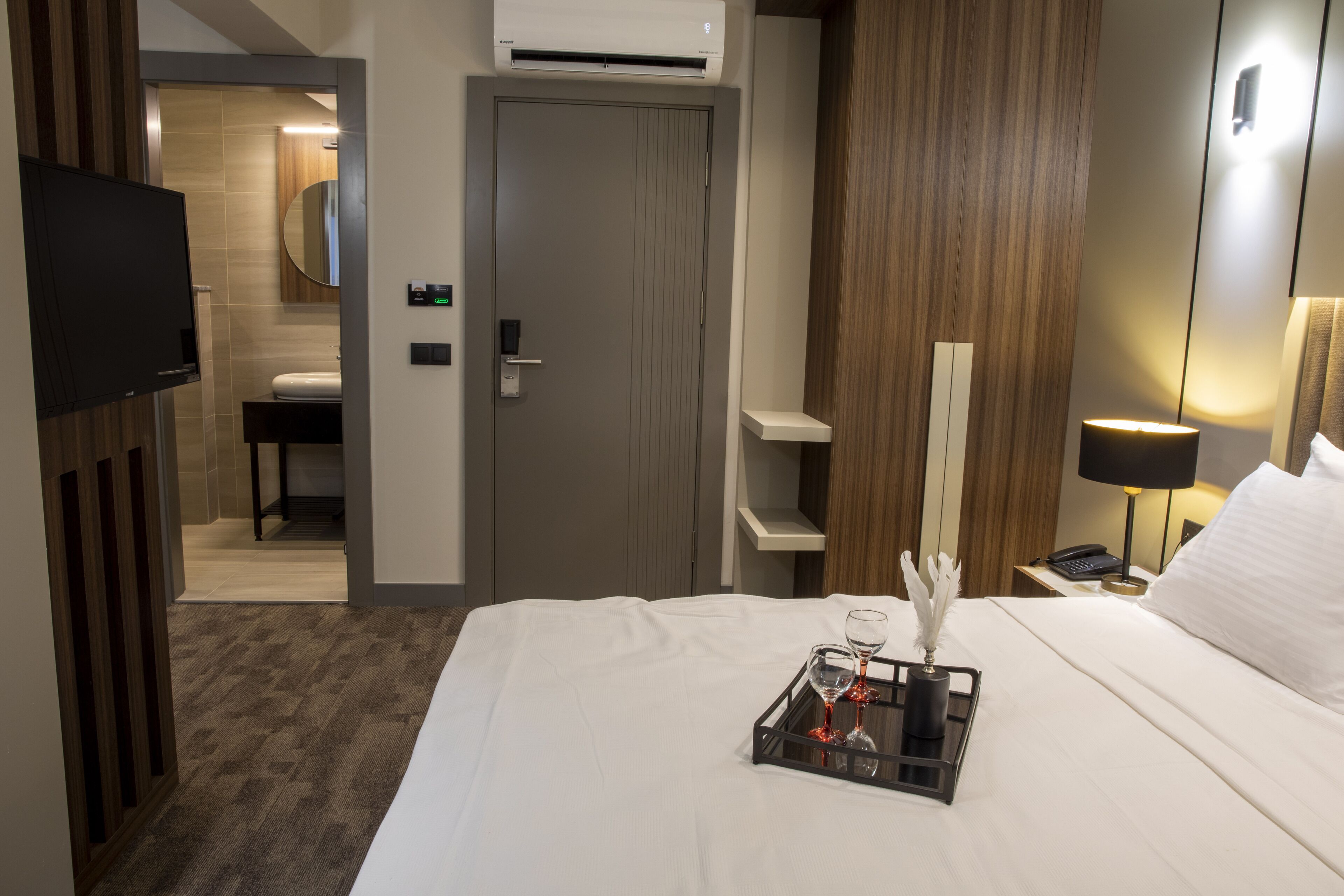 A hotel room with a bed, a bedside table with a lamp, a TV, a bathroom visible through an open door, and a tray with a glass and feathers.