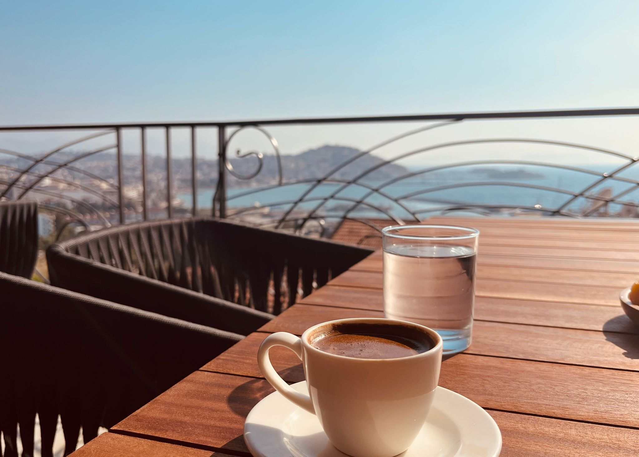 A wooden table with a cup of coffee and a glass of water, overlooking a scenic view of mountains and a body of water.
