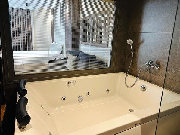 A modern bathroom with a large bathtub, a shower with a handheld hose, and a window with blinds. - Monalisa (Kusadasi)