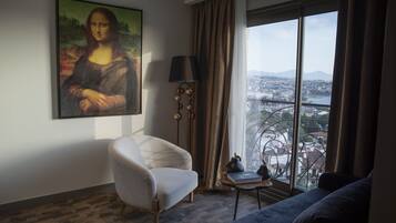 A modern hotel room with a large painting of the Mona Lisa, a comfortable armchair, a sofa, a small table with a lamp, and a view of a cityscape through large windows.