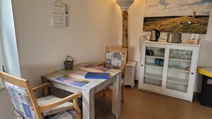A rustic dining area with a wooden table and chairs, a wall-mounted lamp, a cabinet with kitchenware, and a pastoral painting.