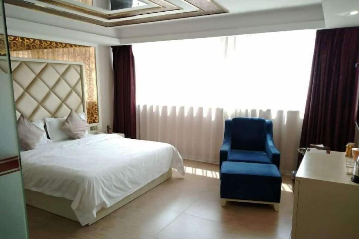 A hotel room with a bed, a blue armchair, a desk, and a window with curtains.