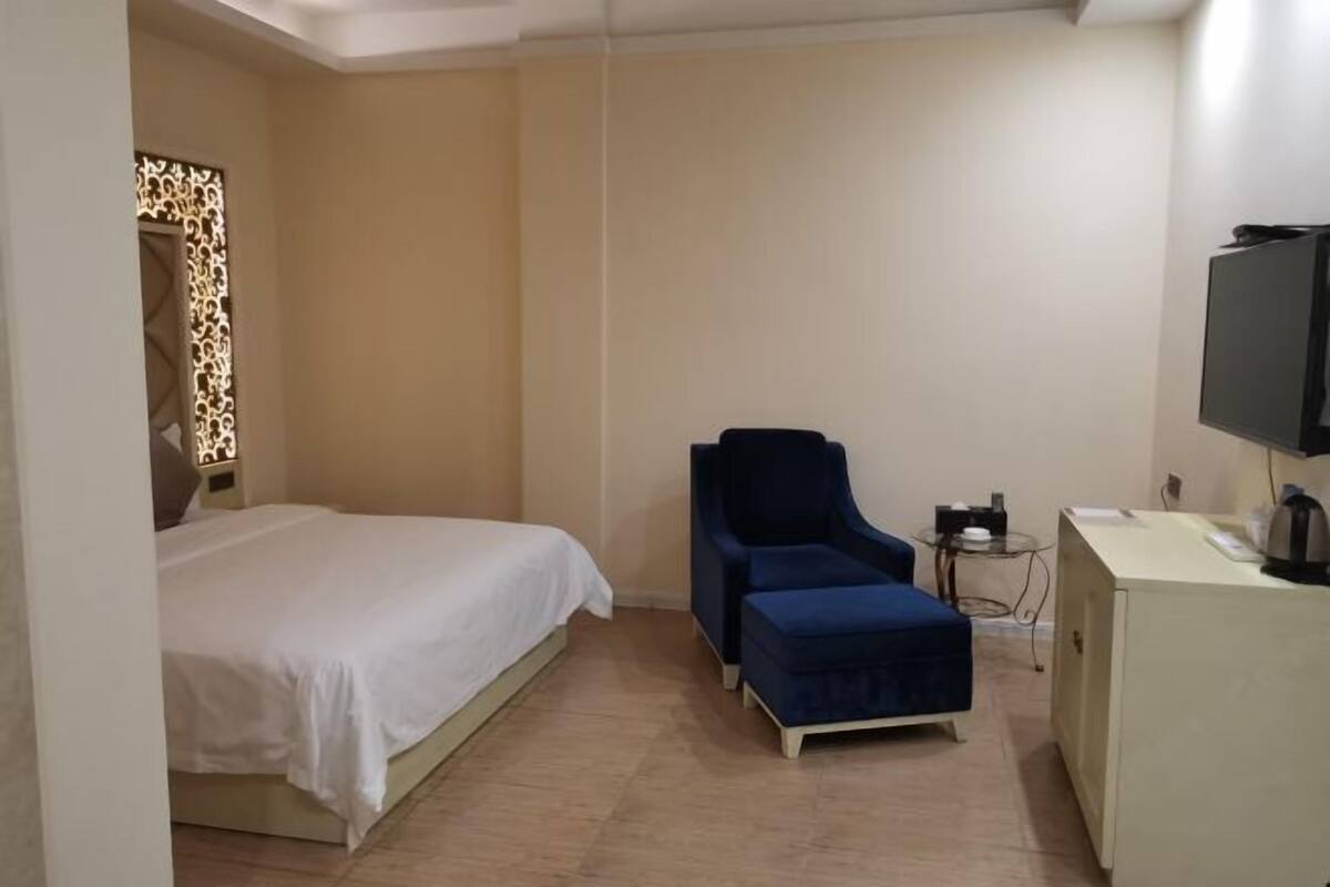 A hotel room with a bed, a blue armchair, a small table with a lamp, and a television on a cabinet.