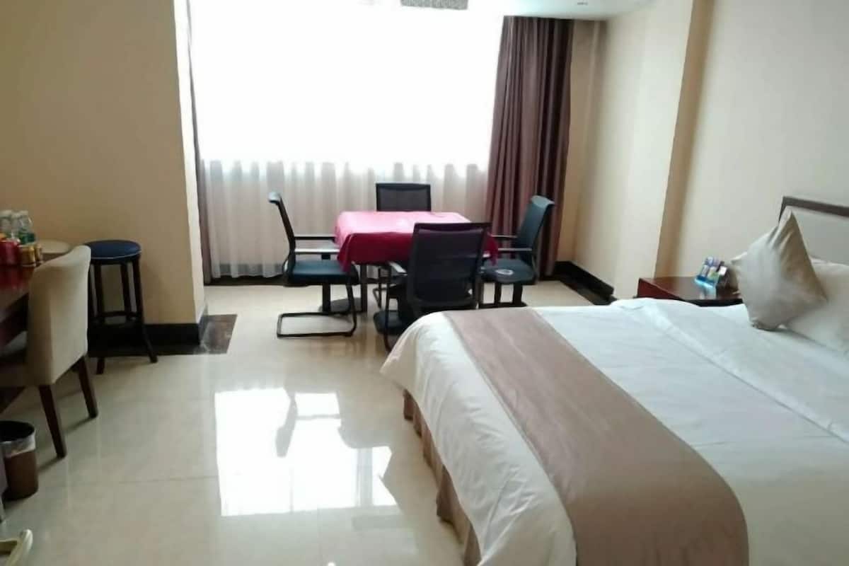 A hotel room with a bed, a desk with a chair, a small table with a red tablecloth, and a window with curtains.