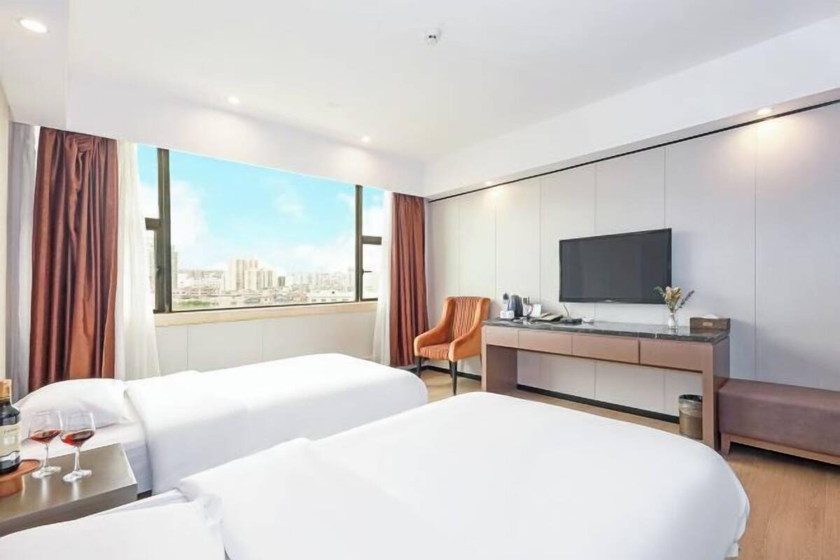 Une chambre d’hôtel avec deux lits, un bureau, une chaise, une télévision et une grande fenêtre donnant sur la ville.