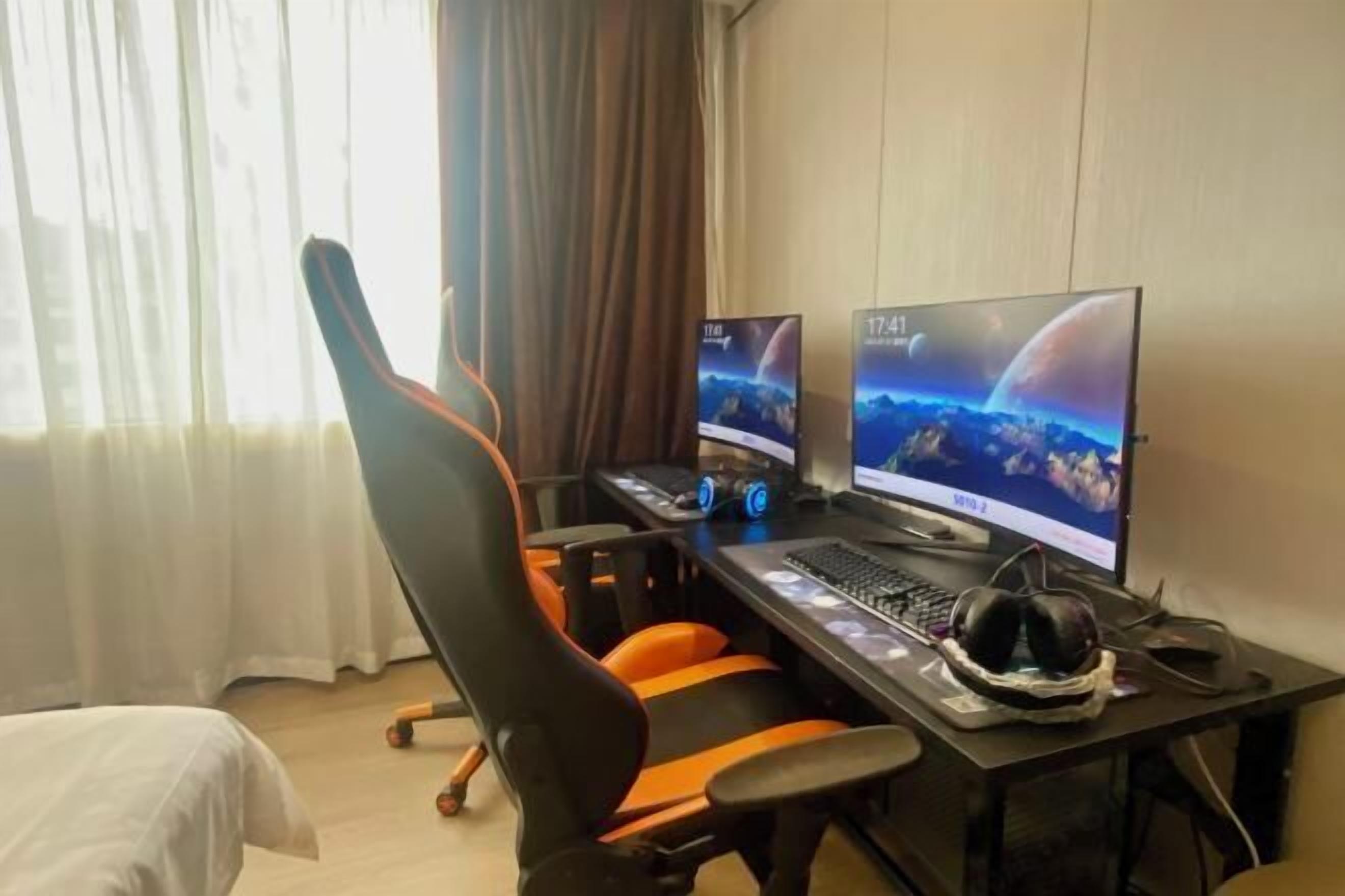 A hotel room with a gaming desk, two monitors, a keyboard, and a gaming chair.