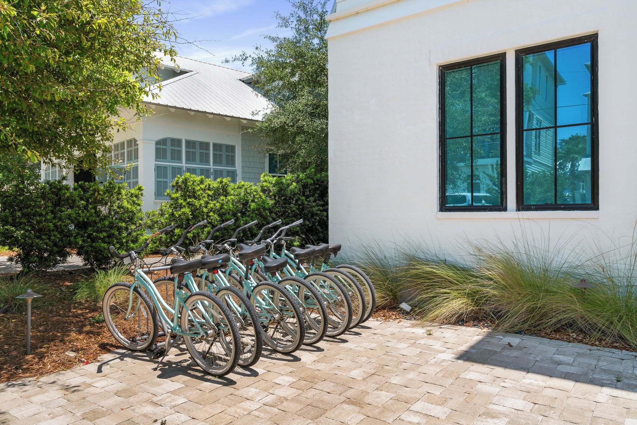 Une rangée de vélos est garée devant un bâtiment doté d’une grande fenêtre, le tout étant entouré de verdure et bordé d’un chemin pavé.