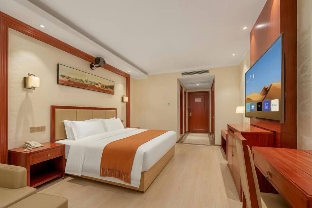 A hotel room with a bed, bedside table, lamp, TV, and a painting on the wall.