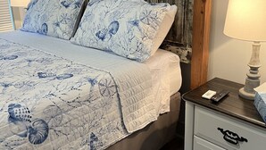 Una cama con una colcha azul estampada con flores, dos almohadas a juego, un cabecero de madera, una mesita de noche con una lámpara y un mueble auxiliar con un control remoto.